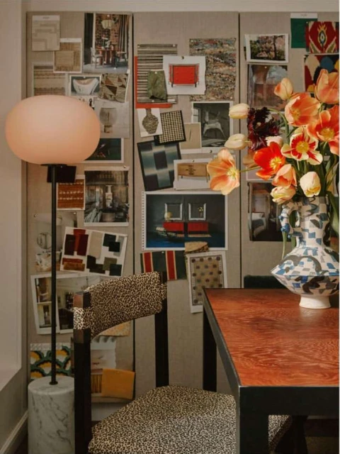 Interior design of a room with a lamp, flowers, table, and a chair with patterned upholstery against a wall with pinned images.