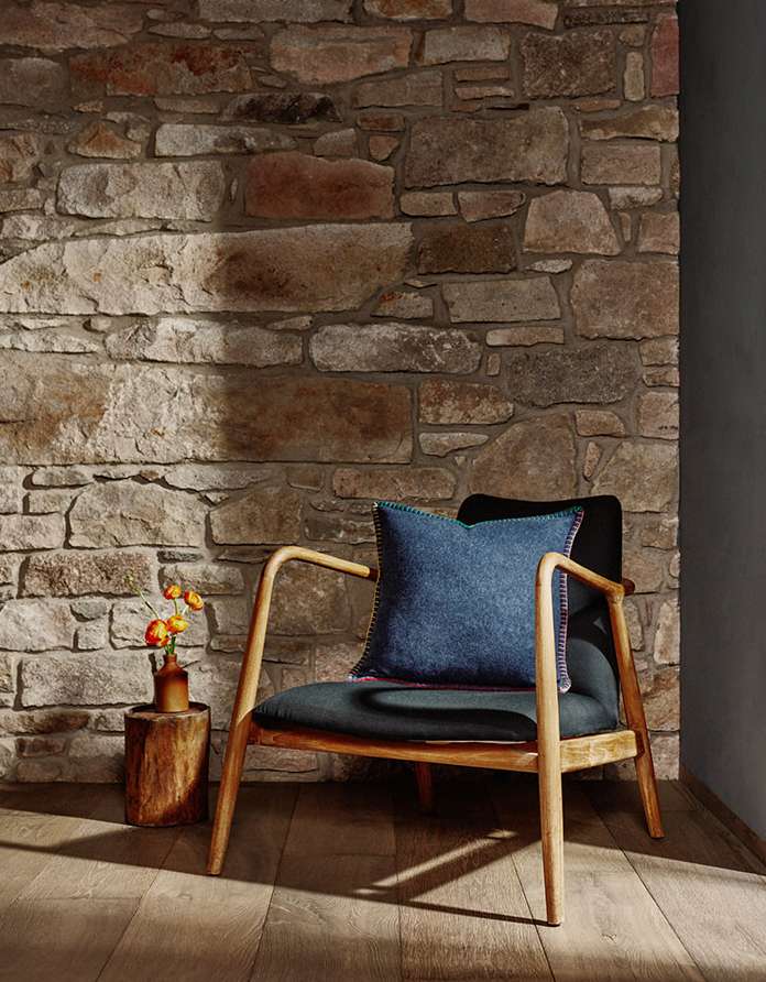 Interior view of wooden chair with cushion and throw pillow alongside a wooden side table with flowers, set against a stone wall.