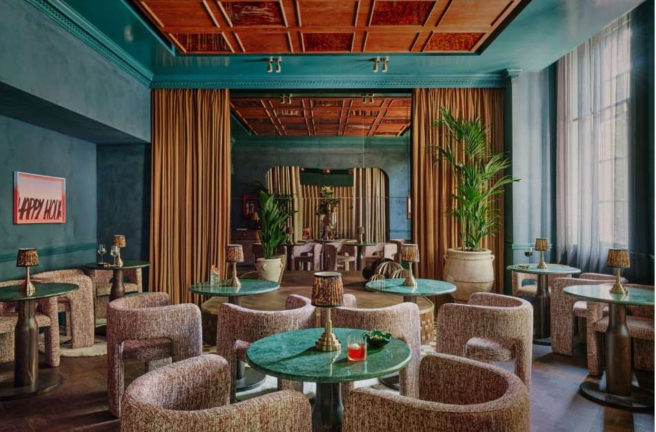 Elegant interior of a stylish bar or lounge area with textured chairs, round tables with green marble tops, potted plants, and dim lighting.