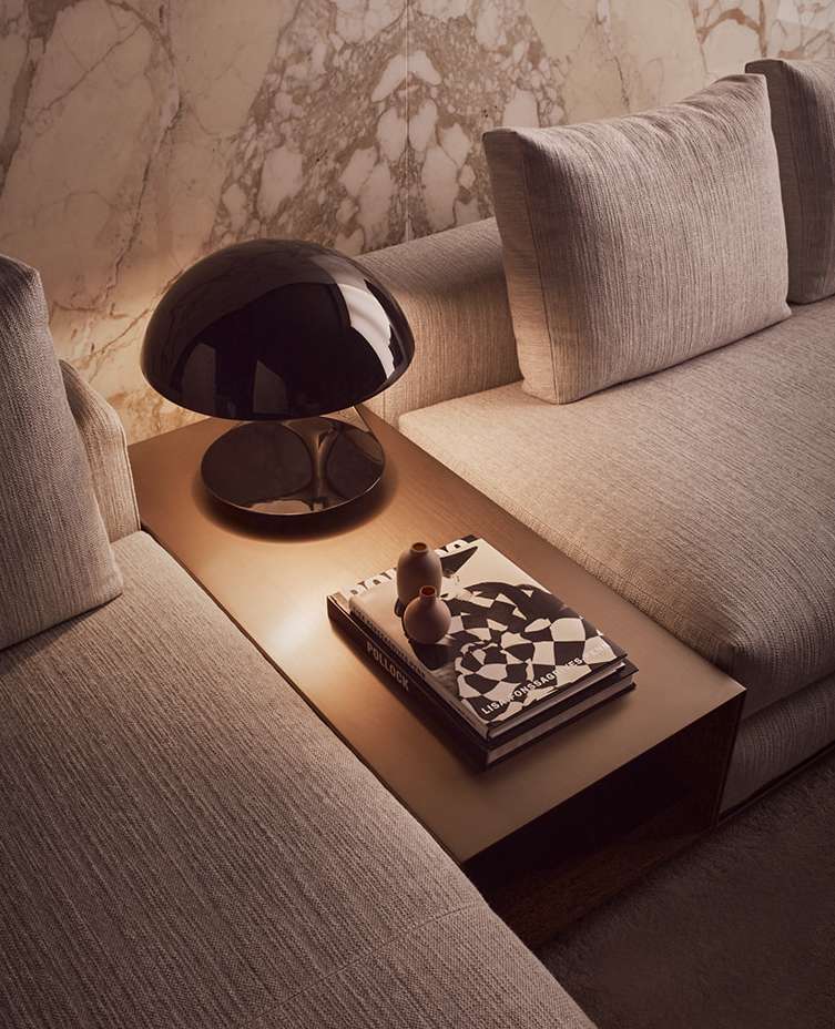 Black mushroom table lamp illuminates books and vases on a side table next to a couch against a marble wall in an indoor setting.