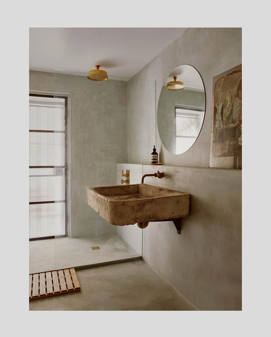 Stone sink and round mirror in a minimalist bathroom with beige walls and a glass-enclosed shower featuring a wooden floor mat.