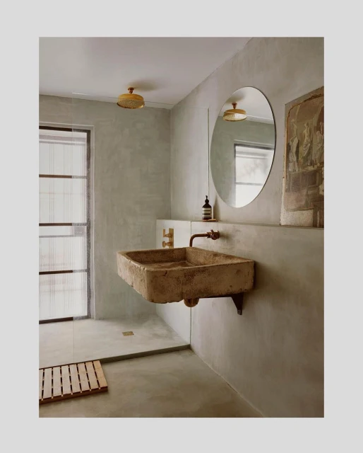 Stone sink and round mirror in a minimalist bathroom with beige walls and a glass-enclosed shower featuring a wooden floor mat.