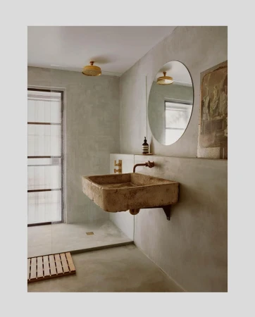 Stone sink and round mirror in a minimalist bathroom with beige walls and a glass-enclosed shower featuring a wooden floor mat.