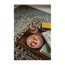 Overhead still life of a rattan tray on an ottoman with madeleines on a patterned plate, a striped mug, a newspaper, and a yellow book.