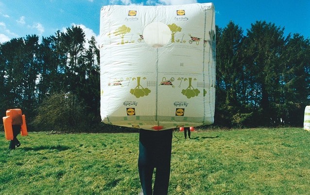 Person in a surreal costume made of a large, patterned white box stands on a green field with trees and blue sky in the background.