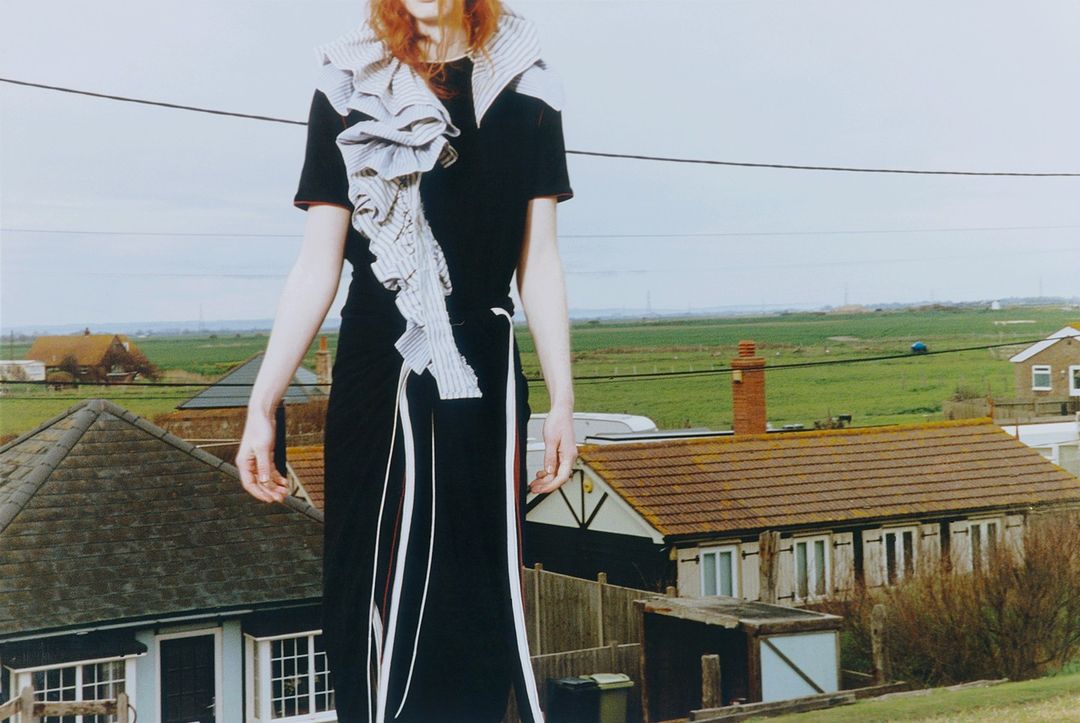 Fashion shot of a woman in a black dress with a white ruffle accent standing outdoors against a backdrop of houses and green fields.