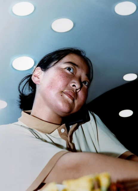 Low angle portrait of an Asian adult female in a polo shirt. Overhead lighting reflects on the face and interior ceiling. Table is visible.