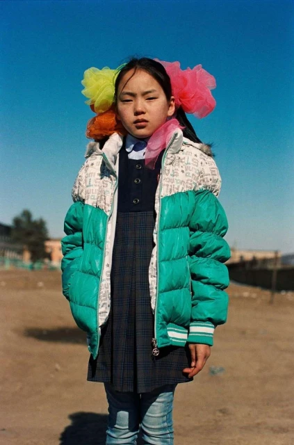 Young Asian girl stands outdoors wearing a green puffer jacket and colorful hair accessories under a clear blue sky. Portrait shot.