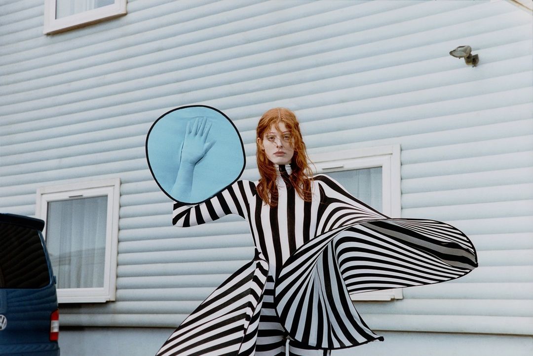An adult woman stands outdoors in a striped dress holding a circular mirror reflecting a hand against a light blue house and a dark blue car.