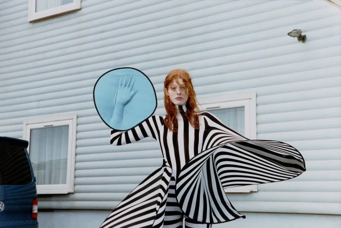 An adult woman stands outdoors in a striped dress holding a circular mirror reflecting a hand against a light blue house and a dark blue car.