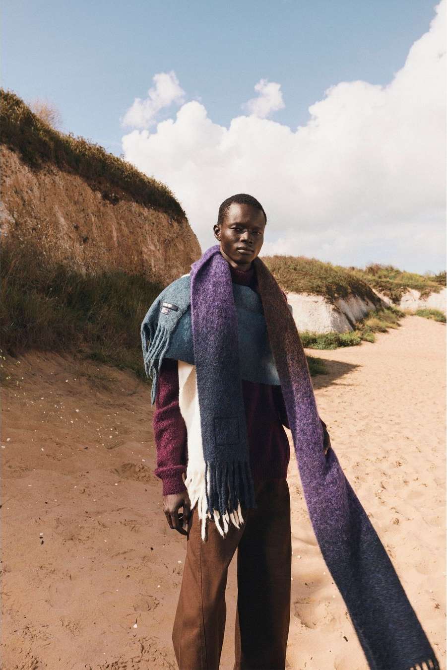 An African male model stands on a sandy beach with cliffs in the background, showcasing a burgundy sweater and long blue-purple scarf.