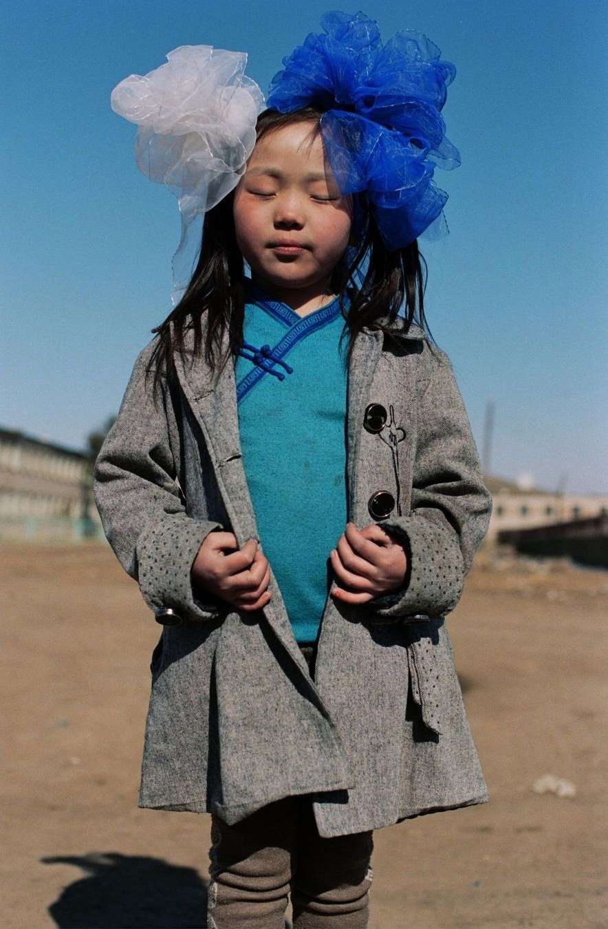 Young girl in a grey coat with blue and white tulle headpiece standing outdoors against a clear sky and tan ground.