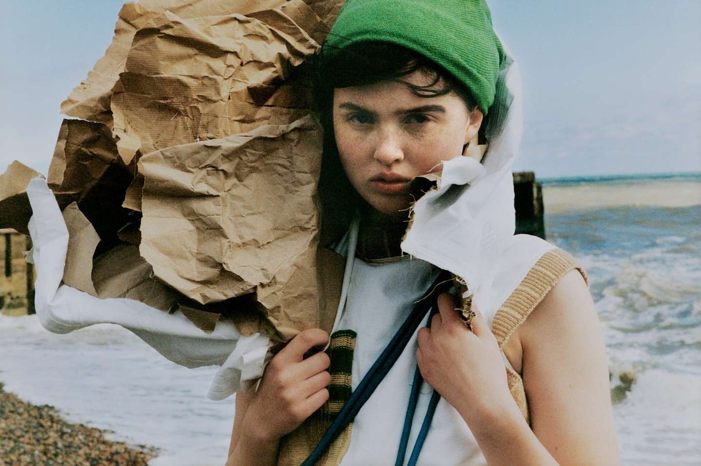 A young woman with a green hat stands on a pebble beach, holding a large bundle of crumpled brown paper, looking directly at the camera.