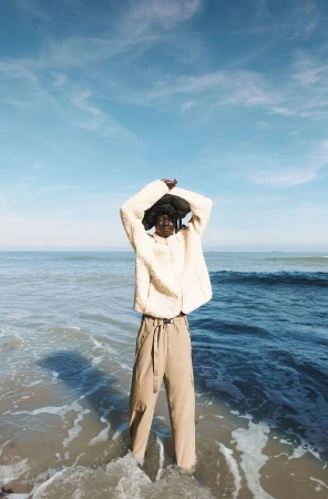 A Black woman stands in the ocean surf on a sunny day, wearing a cream-colored jacket and beige pants, with arms raised overhead.
