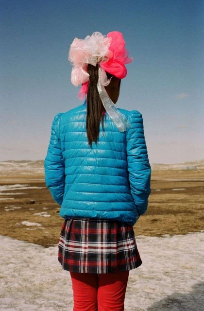 A girl from the back in a field wearing a blue puffer jacket, plaid skirt, red leggings, and a large tulle hairpiece stands outdoors.