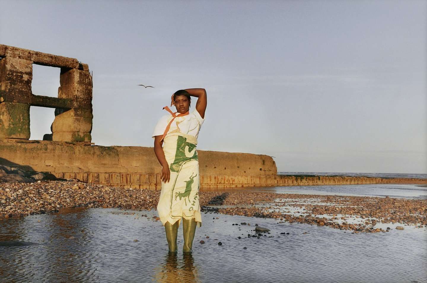 A woman stands in shallow water on a rocky beach near a crumbling stone wall, wearing green boots and a draped white fabric.