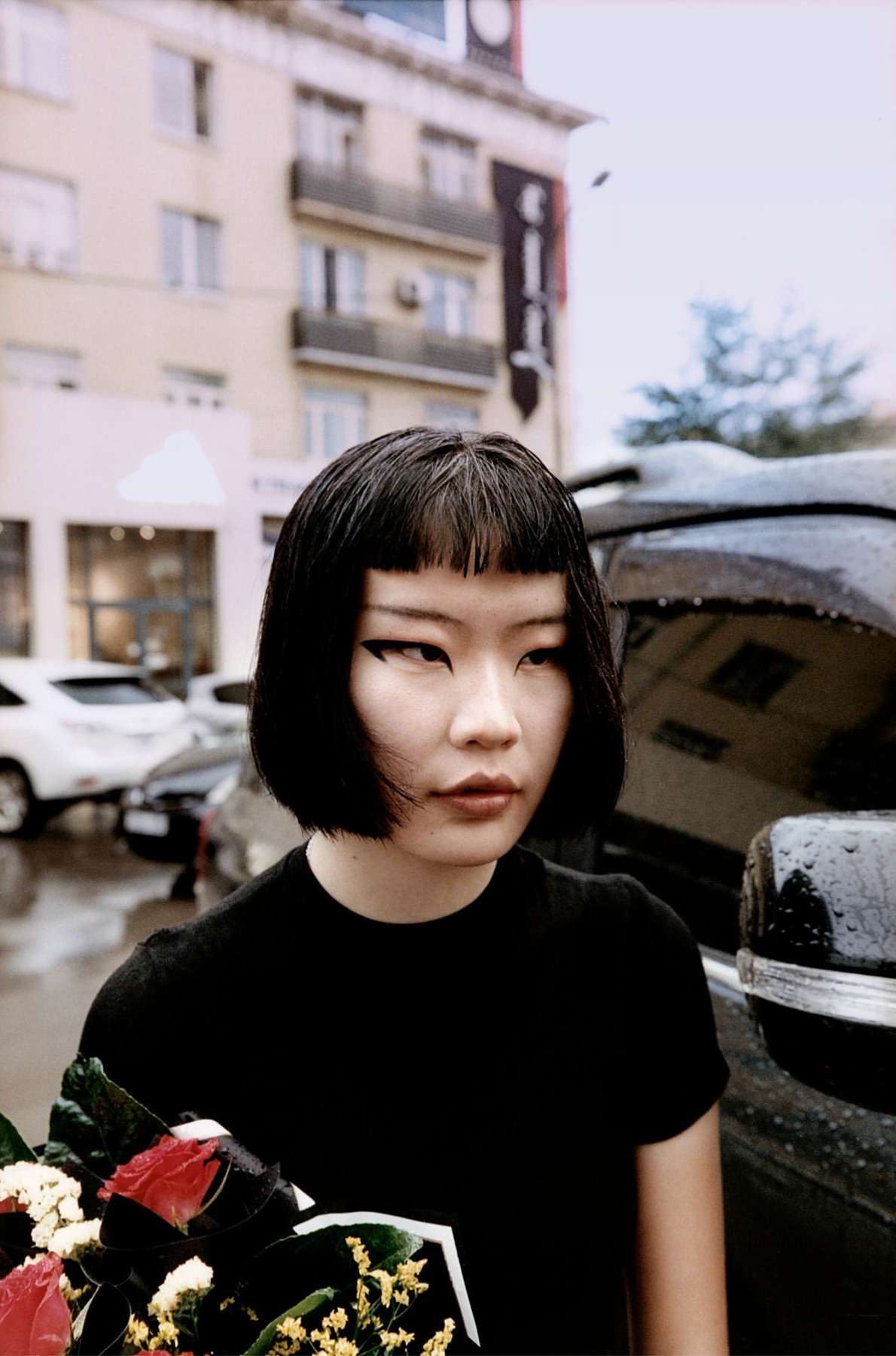 Woman with black hair and winged eyeliner holds a bouquet of red roses and white flowers next to a car in an urban setting.