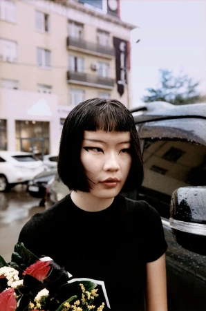 Woman with black hair and winged eyeliner holds a bouquet of red roses and white flowers next to a car in an urban setting.