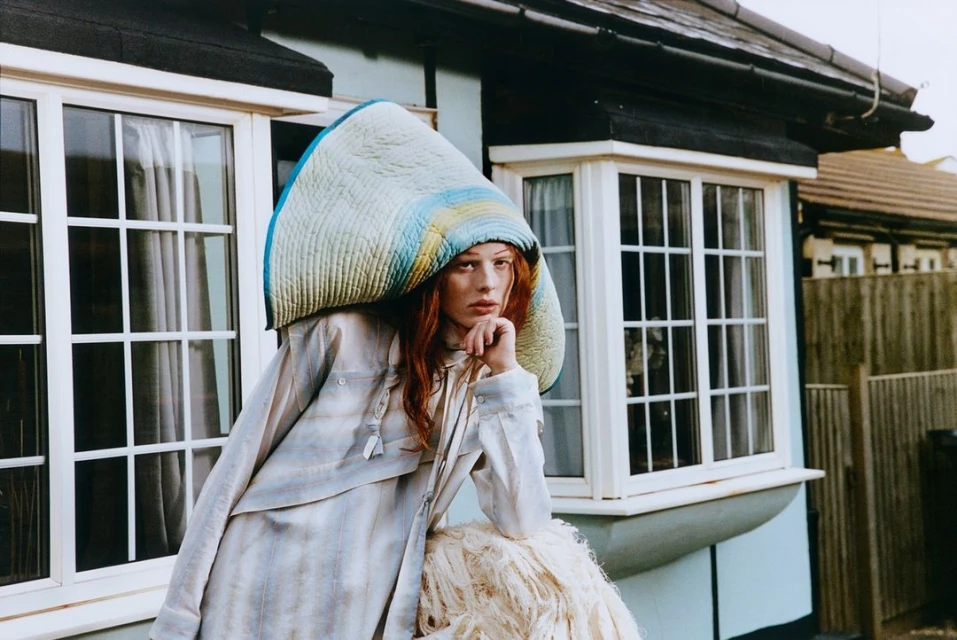 Woman in quilted headpiece and striped shirt leans against a white, shaggy prop in front of a light-blue house with white window frames.