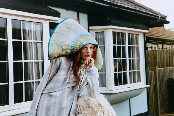 Woman in quilted headpiece and striped shirt leans against a white, shaggy prop in front of a light-blue house with white window frames.