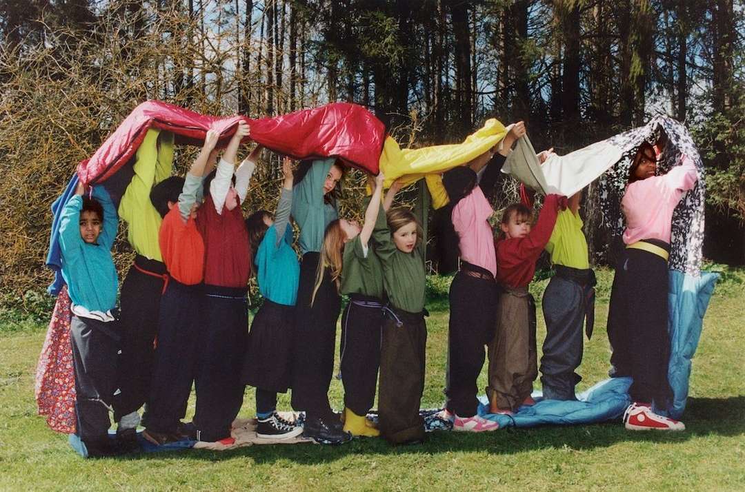 Group of children standing on blue padding while holding up a colorful parachute in an outdoor, grassy environment with trees.