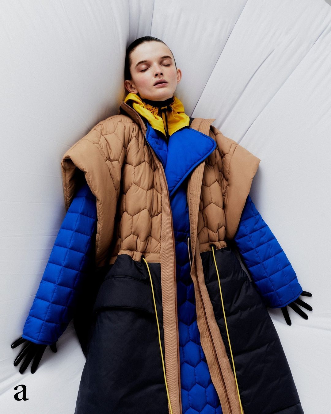 Fashion editorial shot of a stylish woman in a tan, blue, and black puffer coat, laying against a white background, artful studio setting.
