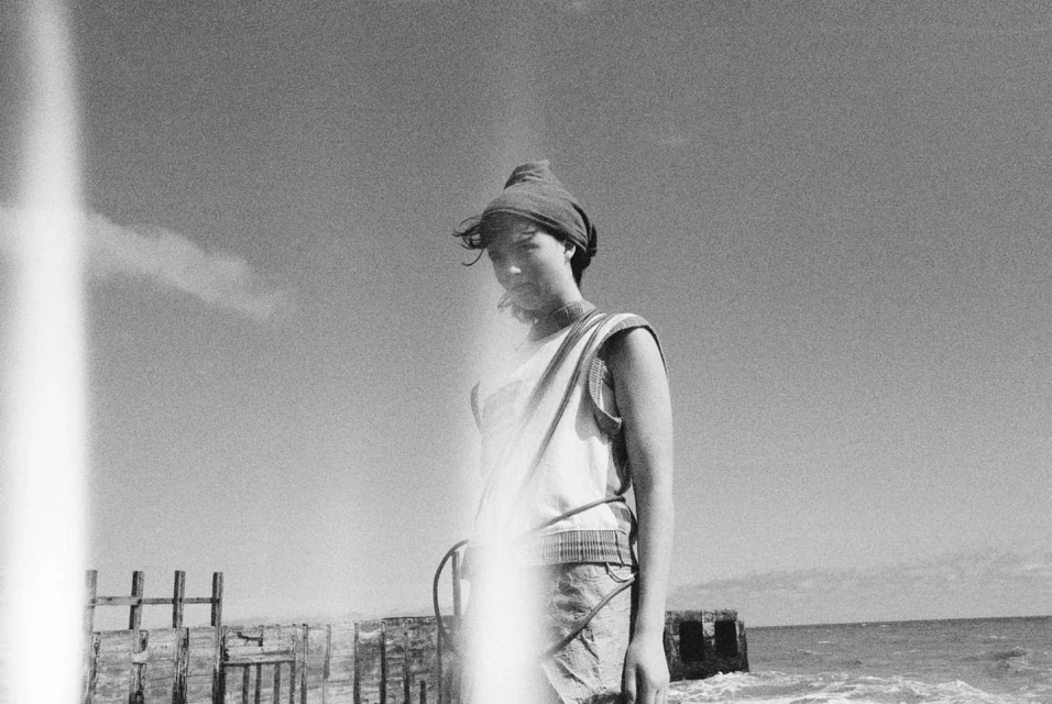 Black and white portrait of a young person in a headscarf standing on a beach with a distant sky and water. Monochromatic.
