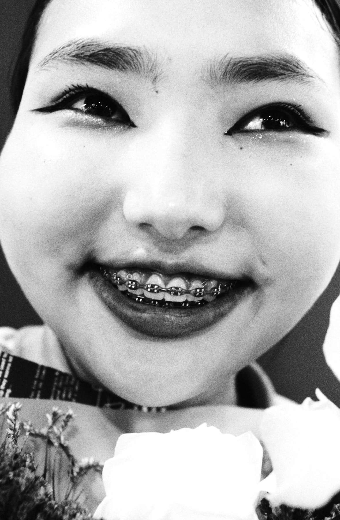 Close-up of a young Asian woman with braces smiling at the camera. Her dark eyeliner is thick and dramatic in a black and white photo.