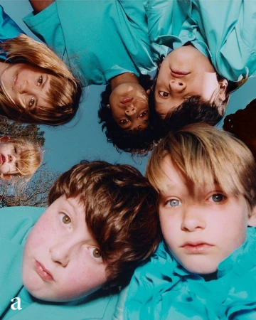 Six young people with light complexions and light brown or blonde hair in matching turquoise shirts pose and look down at the camera.