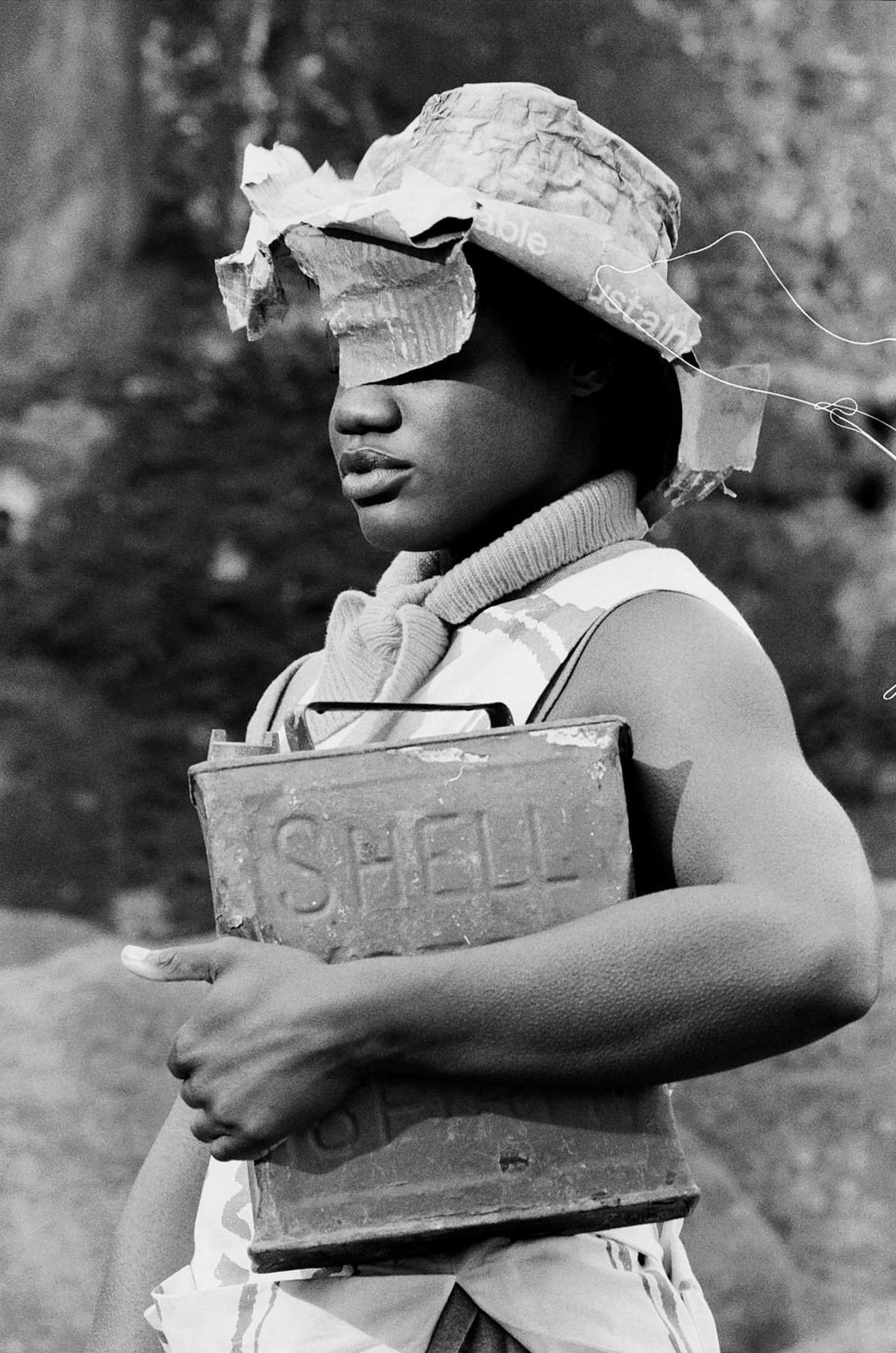 Black and white portrait of a young woman wearing a handmade hat and carrying an old SHELL oil can in an outdoor environment.