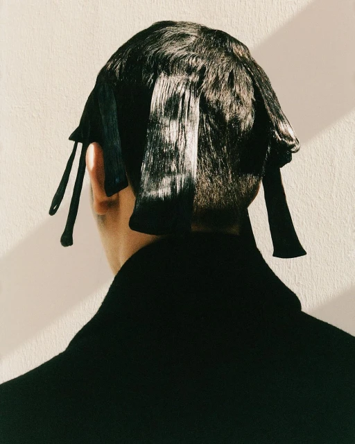 From behind, a person with short dark hair wears an elaborate black hair covering against a plain wall with soft natural lighting.