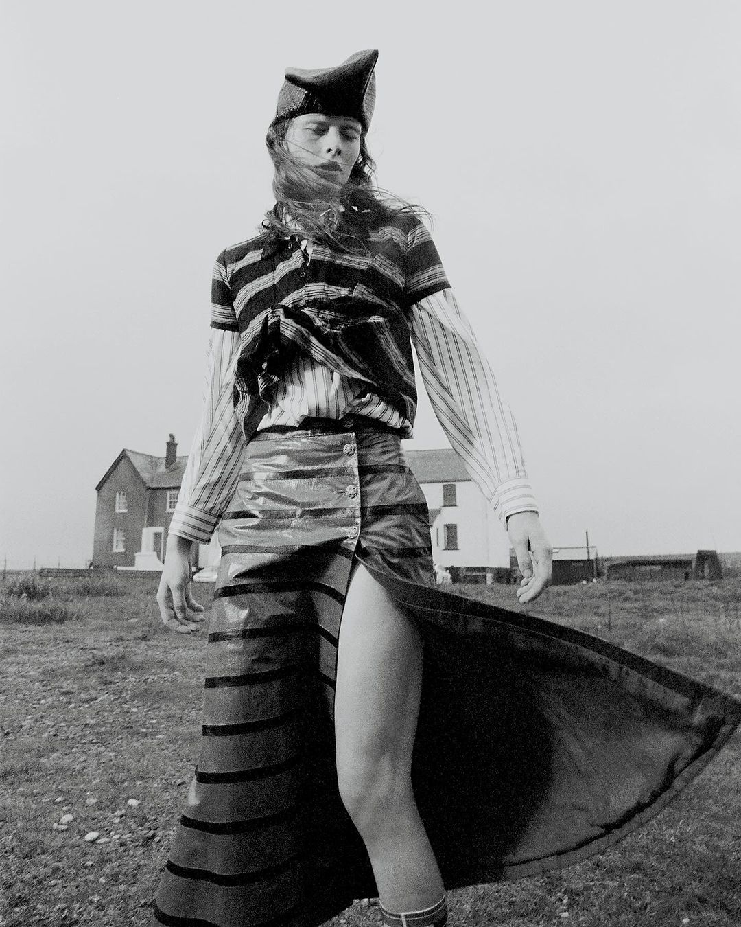 Monochrome fashion shot of a person in striped top, high-slit skirt, and hat posing outdoors with a building in the background.
