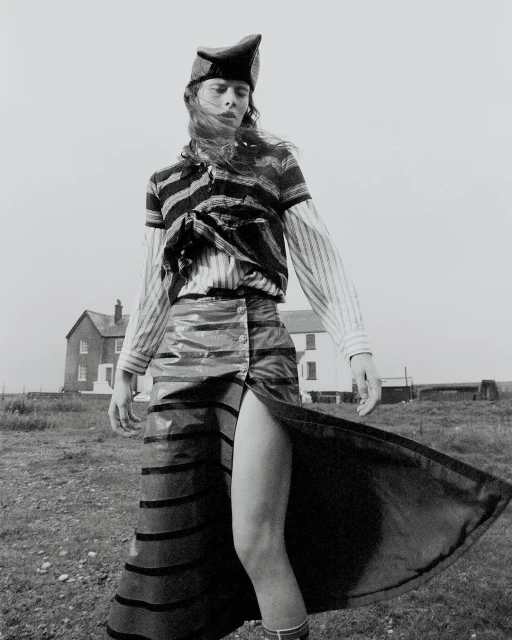 Monochrome fashion shot of a person in striped top, high-slit skirt, and hat posing outdoors with a building in the background.