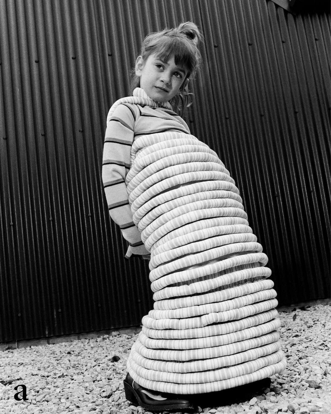 Monochrome portrait of a young girl in a striped top and padded tube-shaped costume standing outdoors with a corrugated metal wall backdrop.