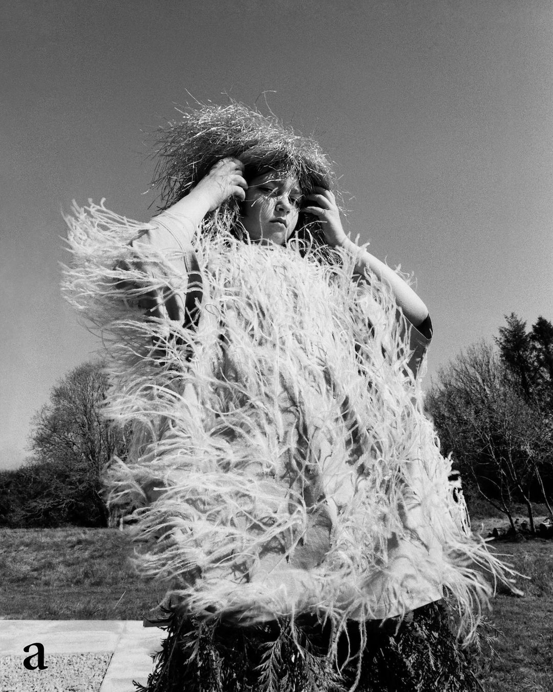 A person stands in an outdoor field, wearing a unique feathered and bristled costume, under a clear sky with trees in the background.