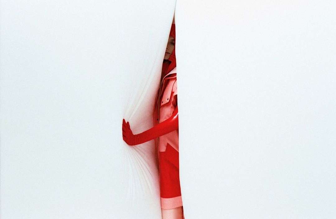 A woman in red gloves and matching outfit peeks through a narrow opening in a white surface, creating a visually striking image.