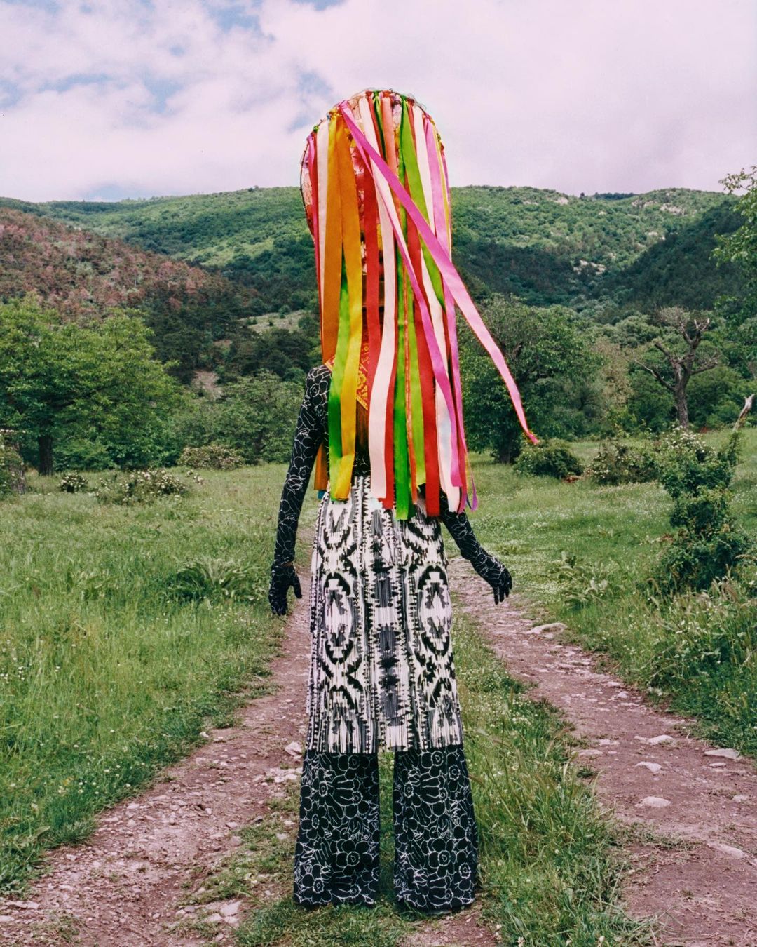 Person with colorful ribbon headdress stands on dirt path in rural landscape with green hills under a cloudy sky.