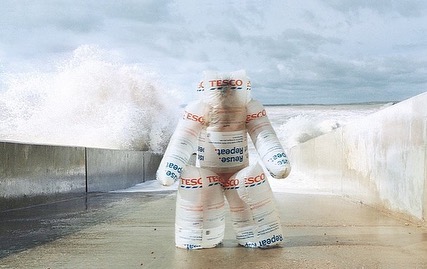 Figure constructed from Tesco brand reusable bags on a concrete walkway near a seawall as waves crash behind on a cloudy day.