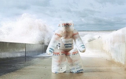 Figure constructed from Tesco brand reusable bags on a concrete walkway near a seawall as waves crash behind on a cloudy day.