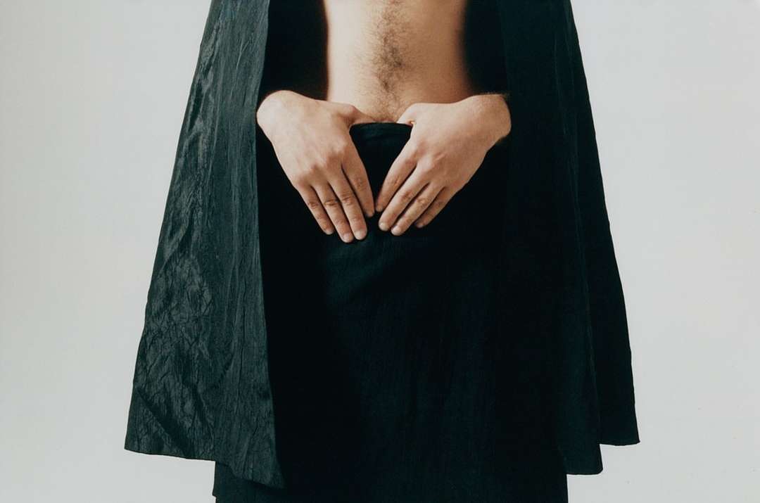 Upper torso of an adult, with their hands clasped over their lower abdomen, partially obscured by black fabric and cape, on white.