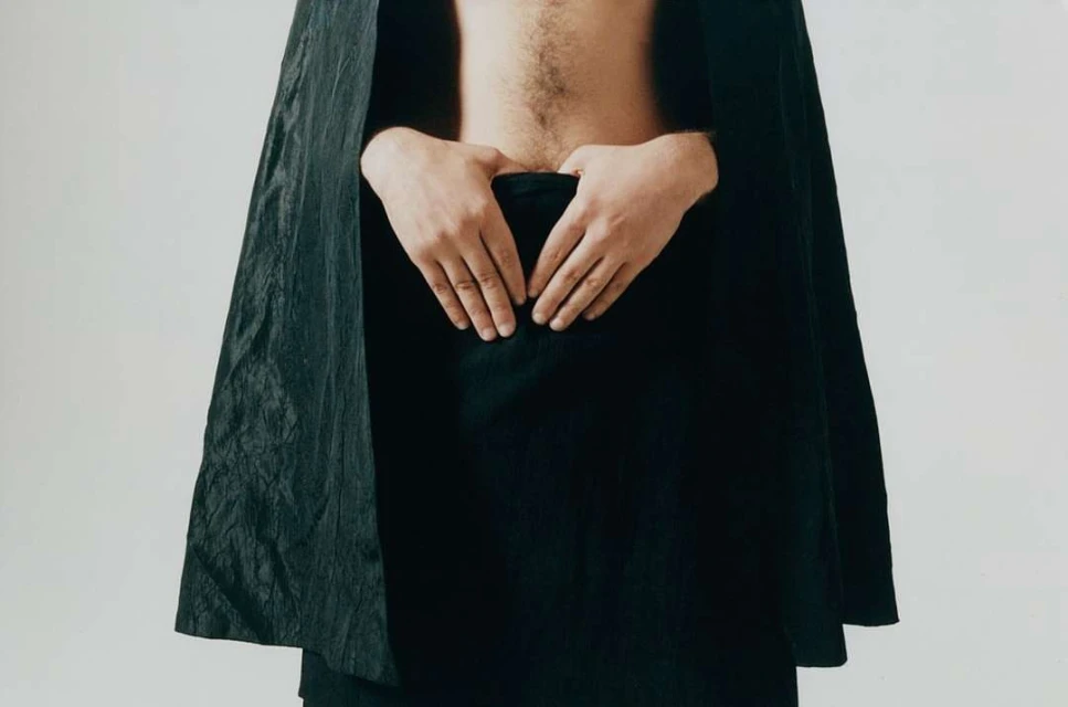 Upper torso of an adult, with their hands clasped over their lower abdomen, partially obscured by black fabric and cape, on white.