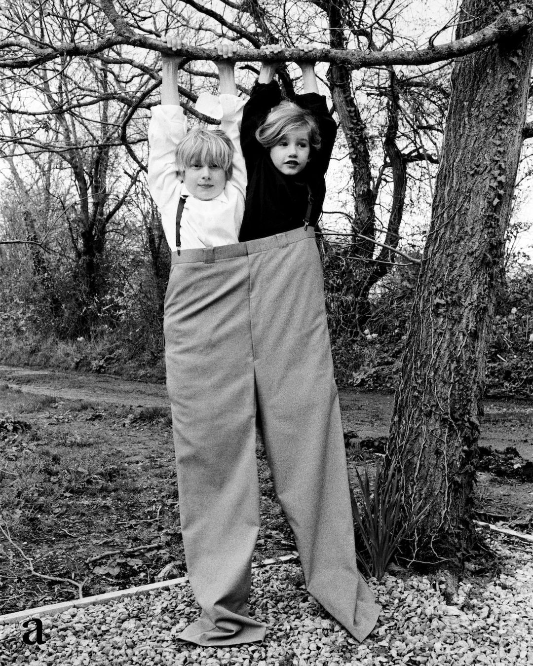 Two children in oversized pants hang from a tree branch outdoors. Black and white image of kids posing playfully in nature.