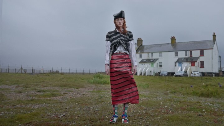 Fashion shot of a redhead woman in an unorthodox outfit posing in a field; a row of white houses is in the background on a cloudy day.