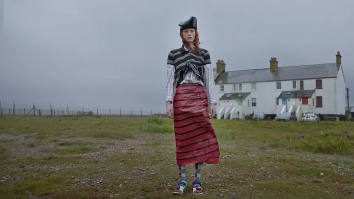 Fashion shot of a redhead woman in an unorthodox outfit posing in a field; a row of white houses is in the background on a cloudy day.