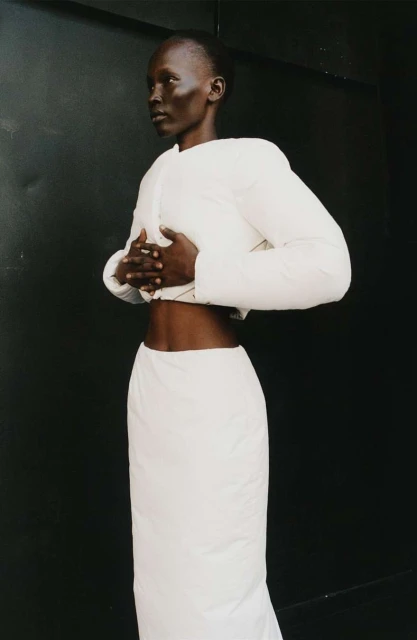 Fashion shot of a Black woman wearing a cream-colored puffy jacket and matching maxi skirt, standing in front of a dark background.