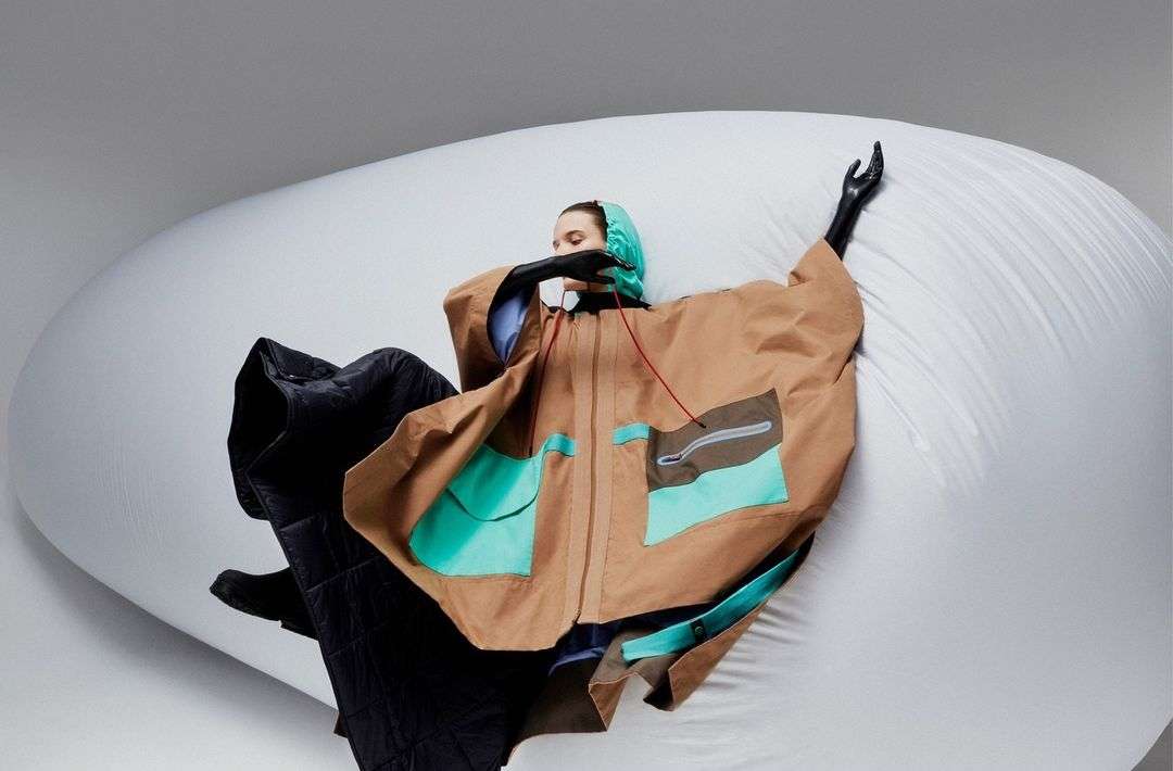 Woman poses in a beige poncho with teal pockets, wearing black gloves and a teal head covering, set against a white backdrop.
