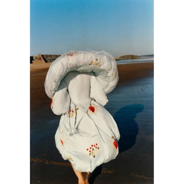 Person shrouded in patterned textile stands on a wet sandy beach under a clear sky, artistic fashion photograph, vertical format.