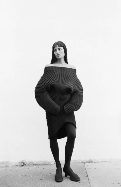 Black and white shot of a woman in a dark dress, tights, and heeled shoes posing against a light background.