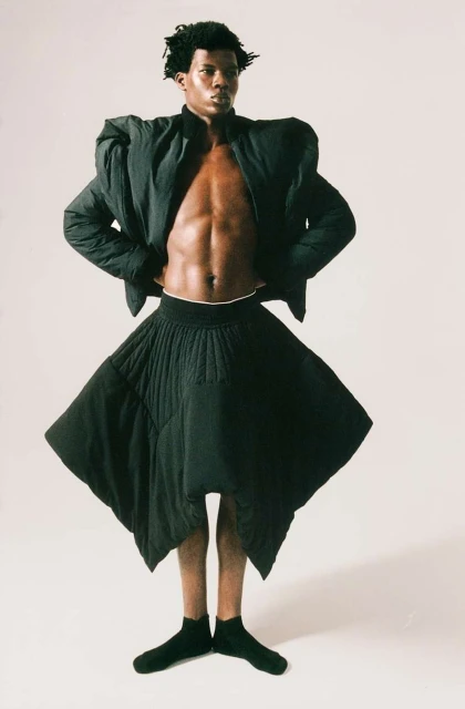 Fashion-forward portrait of one adult male model in a puffy black jacket, black skirt, and black socks posing against a white backdrop.
