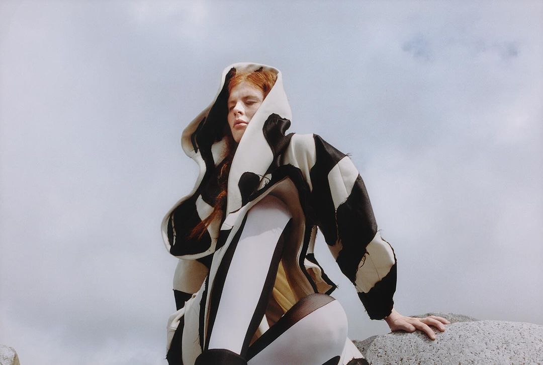 Model with red hair wearing a black and white hooded patterned coat against a cloudy sky